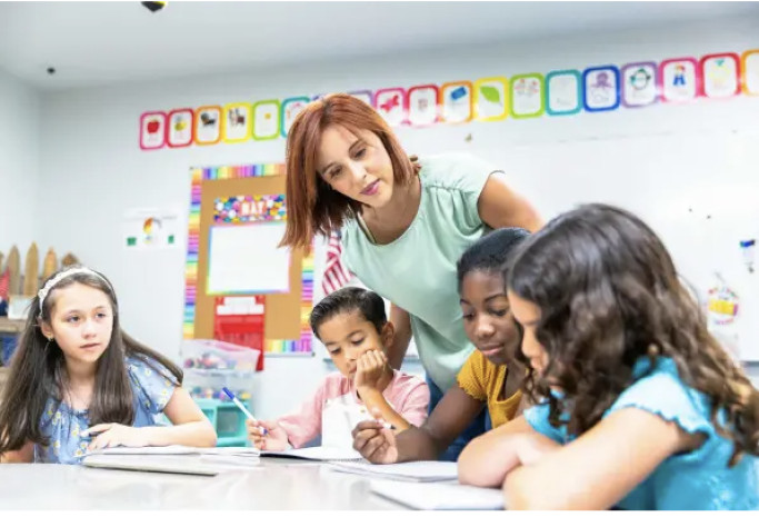 A teacher leans over a table where young children draw in a classroom.